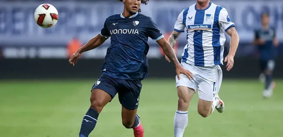 Player in dark kit chasing ball with opponent nearby